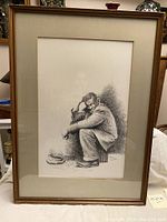 Framed original ink drawing of a man sitting, holding his head, with a hat on the ground in front of him.