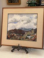 Framed original signed oil painting with landscape theme displayed on an ornate metal stand.