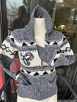 Front and angled view of a gray, cream, and black printed TNA zip-up cardigan sweater on a mannequin showing the hood, front pockets, and toggle zipper pull.