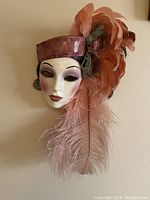 Front view of vintage ceramic porcelain face mask decorated with feathers, showing facial makeup and headband with feather detail.