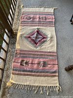 Top-down photo of vintage Mexican style wool rug showing geometric diamond center and striped bands with small stylized designs, light toned fringes on edges.