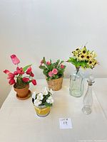 Five faux flower arrangements and two glass bottles displayed on table. Pink tulips in clay pot, pink flowers in basket, white flowers in metal pot, yellow bouquet in green glass bottle, and clear bottle with decorative stopper visible.