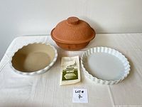 Front view of two fluted white ceramic baking dishes (one shallow, one deep) and terracotta clay roasting pan with lid and recipe book on white background.