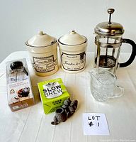 All coffee and tea items arranged on table: two ceramic canisters with lids featuring antique design, Bodum French press coffee pot, glass milk pitcher, tea time thermometer in packaging, loose tea brewer in green box, and a loose tea brewer figurine.