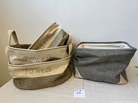 Three fabric storage bins; two beige/tan embroidered with 'towels' and one gray bin with white lining.