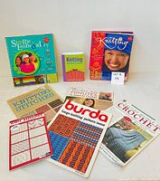 All eight items including embroidery kit, knitting instruction books, crochet pattern book, and kits laid out on white surface