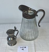 Two pitchers side by side on a white surface. Larger fluted glass pitcher with silver handle and spout, small pewter pitcher with lid and handle.