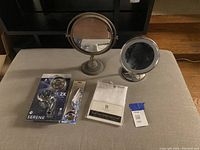 Photo of the two adjustable round makeup mirrors, the Waxman Serene shower head in packaging, shower arm flange, and white fabric shower curtain on a beige surface.