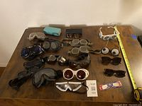 Overhead photo showing 15 goggles and glasses arranged on wooden surface with measuring tape for scale