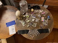 Overhead view of round wooden table displaying lapel pins individually bagged, rhinestone choker on black band, glass jar, egg-shaped gold stand decor, and assorted small decor items.