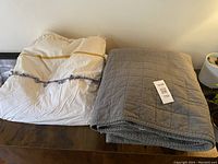 White boho style duvet cover with tassels and decorative trim folded on left; Casaluna gray quilt with tag folded on right on wood surface.