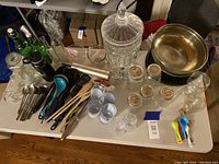 Photo of assorted kitchen items including glass jars, metal bowls, a crystal glass beverage dispenser, various utensils, measuring spoons, small labeled jars, and glasses on a table.