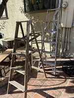 Two metal ladders and one wooden ladder positioned outdoors on brick floor against wall.