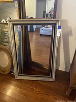 Large rectangular mirror with ornate vintage silver frame leaning against a wall on wood floor. The frame shows decorative molding and a muted silver finish.