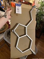 Silver metal serotonin molecular wall art lying on a cardboard surface with hexagonal and pentagonal rings clearly visible, hand holding the art in one photo.