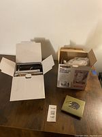 View of all three items side by side on wooden table: open box with pasta maker, open box of snow cone machine, ceramic trivet with badger design.