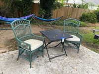 Full set showing two green wicker chairs with cushions and the black folding metal table outdoors on stone patio with some leaves on the ground and table.