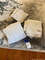 Three folded beige sheets with subtle print, arranged on a patterned rug. A printed card with number 100 is also visible.