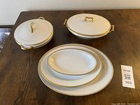 Set of six white porcelain serving dishes with gold rims and gold handles on covered dishes, displayed on a wooden table.