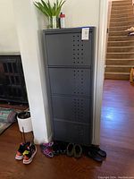 Full view of closed grey metal shoe storage cabinet showing four front-hinged compartments and ventilation holes, placed against the wall with various shoes on the floor around it.