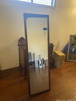 Full front view of a tall rectangular standing mirror with a dark wooden frame, reflecting part of the room including furniture and boxes.