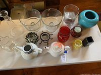 Image shows two white ceramic teapots, large clear glass trifle bowls (round and square), various colorful vases, and a clear glass pitcher.
