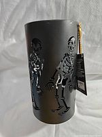 Black cylindrical metal lantern with dancing skeleton cut-out designs and gold handle, side view showing skeleton details.