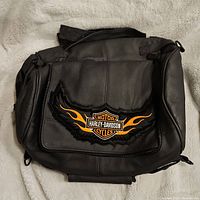 Black leather bag with Harley Davidson logo and orange flame design on front flap pocket.