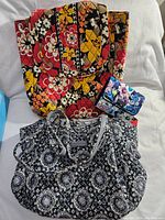 Two Vera Bradley items: black and white patterned purse with silver straps and small blue floral wallet in front of colorful floral backpack