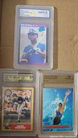 Photo of three graded baseball and sports cards: Ken Griffey Jr. rookie card with WCG grade 10, Greg Maddux 1992 Topps Traded card graded BGS 9.0, and a 1995 Fleer Ultra Swimsuit card graded 10.