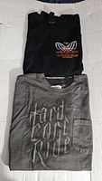 Two folded T-shirts on white surface, top black with chest logo, bottom gray with front pocket and 'Hard Core Rider' text