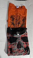 Folded orange Harley Davidson T-shirt with black winged emblem and circular logo, stacked on folded black T-shirt with large skull graphic in red and grey.