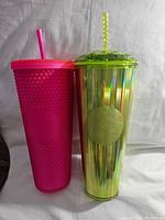Photo of two Starbucks tumblers side by side, one hot pink with textured surface and straw, one iridescent green with smooth reflective surface and straw.
