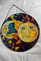 Full view of the round stained glass wall hanging depicting sun and crescent moon faces with rich colors including yellow, blue, orange, and purple. The artwork is framed in metal and has an attached hanging chain.