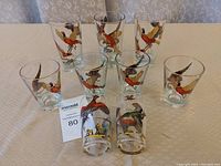 Nine glasses arranged on a white cloth, 6 small and 3 large, all decorated with colorful bird (pheasant) motifs.