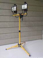 Full view of the yellow adjustable tripod stand with dual halogen work light fixtures mounted on top, set outdoors against siding.