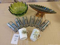 Full view of lot on table including green glass bowl, 6 retro glasses with geometric design, 2 coffee mugs with floral designs, green and gold decorative vase bowl.