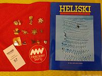 Book titled 'Heliski: The Cariboo & Monashees' with 13 assorted ski pins and Helicopter Skiing sticker on a red fabric background