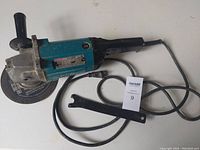 Makita GA7910 angle grinder with grinding disc and changing wrench laid on table showing side profile and attached cord.