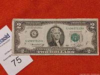 Front of 1976 US $2 bill with serial G49857519A on a red background with auction and lot number cards