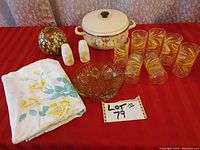 Full kitchen lot including enamel-coated casserole pot with floral design, salt and pepper shakers with yellow flowers, seven clear juice glasses with gold wheat pattern, amber-colored glass bowl, ceramic flower vase with colorful glaze, and folded floral tablecloth on red background.