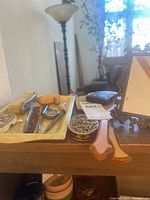 Photo showing assortment of antique hair accessories arranged on a tray and wooden surface including hair combs, powder compact, ornate vanity mirror, decorative pins, and enamelled tin box.