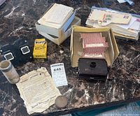 Photo showing a black metal tattooing device, instruction sheet from National Rabbit Supply Company, small containers, 9 music rolls, and a stack of old letters and envelopes