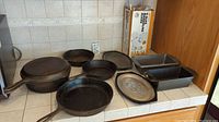 Overview photo showing all cast iron skillets of various sizes, two baking pans, mandoline slicer box, and plastic container with additional items on tiled countertop.