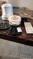 Photo showing all appliances and serving plates: ice cream maker, dessert maker, pizzelle maker, plastic and metal molds, on a marble tabletop.