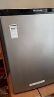 Front view of small Frigidaire refrigerator freezer with a silver door and black trim on top. The appliance has a MaxSold sticker on it.