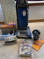 Wide view of Hoover upright vacuum, Dirt Devil vacuum hose and bags, Rowenta iron and accessories arranged on floor.