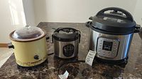 Photo showing three kitchen appliances on a counter: yellow Rival Crockpot slow cooker, Aroma digital rice cooker, and stainless steel Instant Pot electric pressure cooker