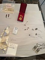 Overview photo showing multiple pins, a white ribbon labeled 'Tunis 1938', a decorative pendant in red velvet box, and smaller pins and ribbons laid out on paper with handwritten notes.