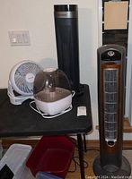 Four small appliances: GiveBest tower heater, Honeywell fan, DreamEgg humidifier on a black table, and woodgrain panel tower heater standing beside the table.